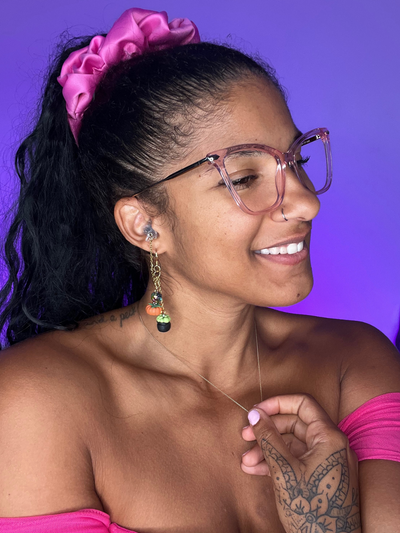 Purple Background. Black woman facing the right wearing high fidelity ear plug earrings in gold with clay charms dangling