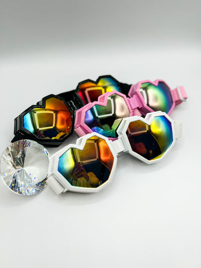 There is a white background. There are three pair of the LoveGaze rave goggles in frame. One of each colour, Black, Pink, and white. beside them is a giant diamond shaped crystal reflecting some light. The goggles are rainbow chrome mirrored lenses.