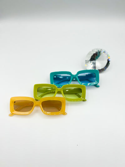 There is a white background with Three pairs of our PopBlocks sunglasses. sitting behind all 3 is a giant diamond shaped crystal refracting light. from top to bottom is our Blue, then Green, Then yellow.