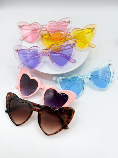 6 pairs of heart shaped sunglasses on a white background. from the back to the front is pastel pink, yellow, purple, and blue. Then pink with black gradient lenses and tortise shell infront with a brown lense.