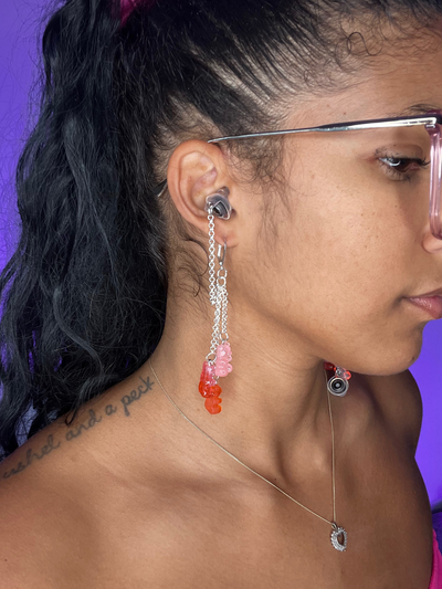 Purple Background. Black woman facing the right wearing high fidelity ear plug earrings in silver with gummy bearsdangling