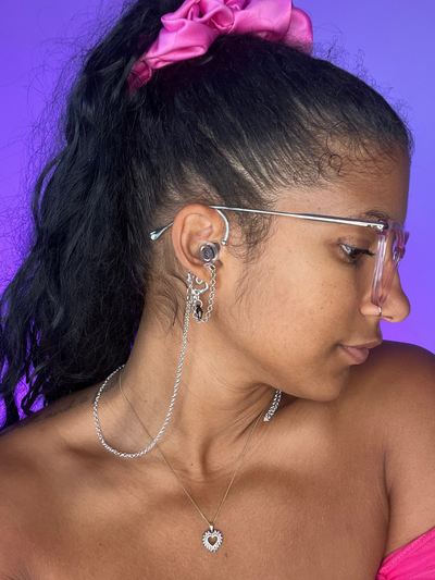 Purple Background. Black woman facing the right wearing over the ear high fidelity ear plug earrings in silver with a Black bead.