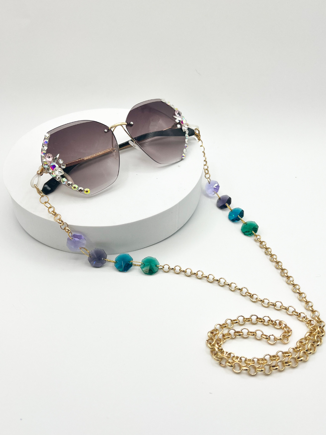 Product shot of Gold rolo chain Sauce Strap Glasses chains, with four beads. light purple, dark purple, blue teal, and green. being worn on large framelesssunglasses that have rhinestones on the sides. a white background.