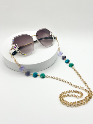 Product shot of Gold rolo chain Sauce Strap Glasses chains, with four beads. light purple, dark purple, blue teal, and green. being worn on large framelesssunglasses that have rhinestones on the sides. a white background.