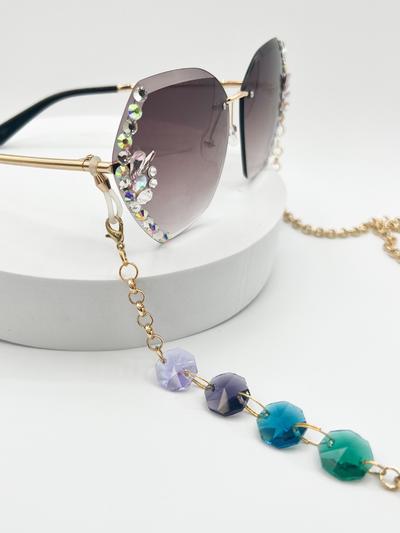 Product shot of Gold rolo chain Sauce Strap Glasses chains, with four beads. light purple, dark purple, blue teal, and green. being worn on large framelesssunglasses that have rhinestones on the sides. a white background.