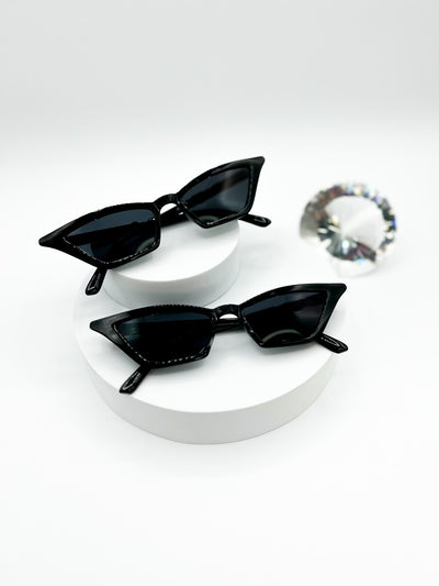 Two pairs of black cat-eye sunglasses with black lenses displayed on white circle risers against a white background. The pointy cat-eye frame adds a stylish touch to this classic design. These sunglasses are available on our website for purchase.