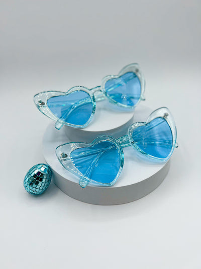 There is a white background with two circular risers stacked ontop of eachother. there are two pairs of identical sunglasses. they are Pastel Blue heart shaped sunglasses with matching lenses. the frames of the sunglasses are transparent