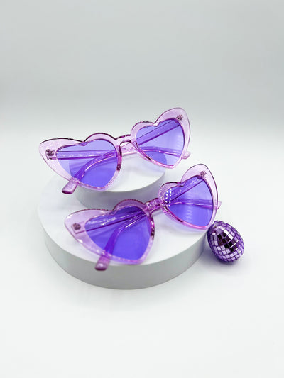 There is a white background with two circular risers stacked ontop of eachother. there are two pairs of identical sunglasses. they are Pastel Purple heart shaped sunglasses with matching lenses. the frames of the sunglasses are transparent