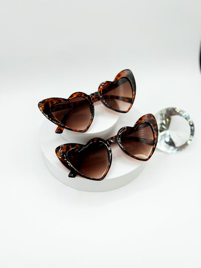 There is a white background with two circular risers stacked ontop of eachother. there are two pairs of identical sunglasses. they are tortoiseshell heart shaped sunglasses with a gradient brown lenses. the frames of the sunglasses are tortoiseshell..
