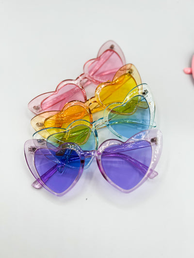 a white background with four pairs of pastel heart shaped sunglasses stacked ontop of eachother. There is Pink, Yellow, Blue, and Purple.