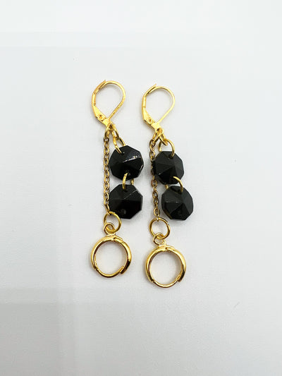  a pair of gold leverback earrings featuring two black glass beads dangling from the post, with a chain and circular earring post at the base are laying flat on a white background.