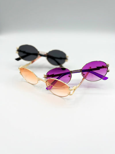 There is a white background with three pairs of sunglasses displayed. all three are the Midnight Gold Style. all three are made with gold hardware. Gold rim and circle lenses. there is Black, Purple, and Peach coloured lenses. the arms of the sunglasses have a zig zag in them and look kind of like a lightening bolt..