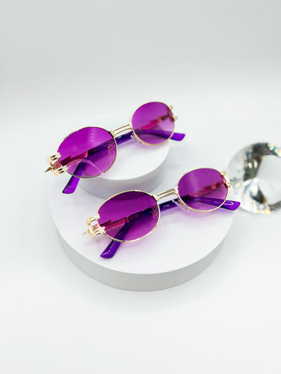 There is a white background and two pairs of our Midnight Gold sunglasses are displayed. Gold rim and circle lenses. there is Purple coloured lenses. the arms of the sunglasses have a zig zag in them and look kind of like a lightening bolt..