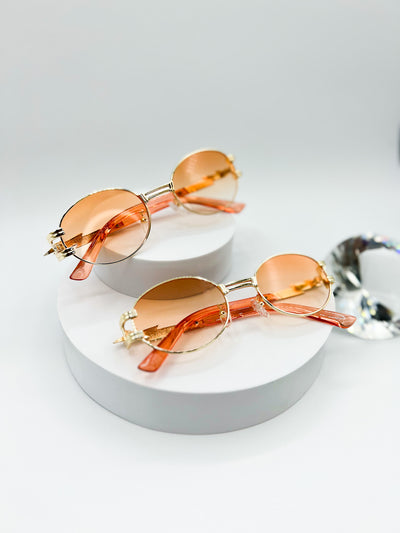 There is a white background and two pairs of our Midnight Gold sunglasses are displayed. Gold rim and circle lenses. there is Peach coloured lenses. the arms of the sunglasses have a zig zag in them and look kind of like a lightening bolt..