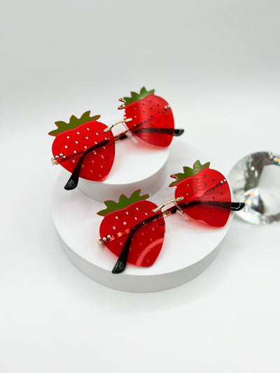A white Background with two pair of the same style of sunglasses shaped like strawberries, rimless, and to simulate all the little seeds strawberries have, there are tiny holes all over the lenses. These lenses are red with little green sprouts on top.