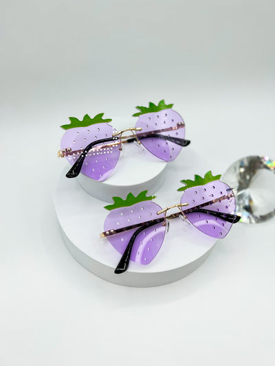 A white Background with two pair of the same style of sunglasses shaped like strawberries, rimless, and to simulate all the little seeds strawberries have, there are tiny holes all over the lenses. These lenses are Purple with little green sprouts on top.