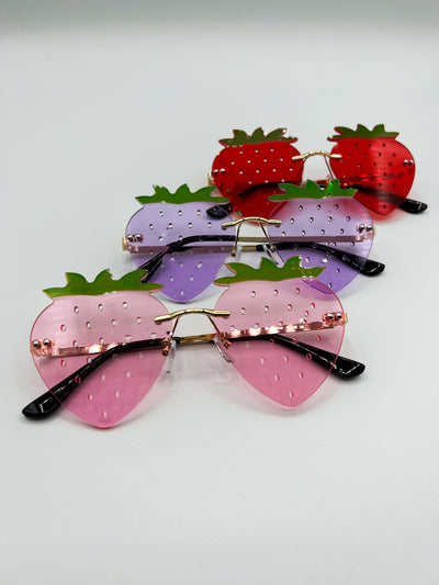 A whiste Background with three pair of the same style of sunglasses in three different colours. They are all shaped like strawberries, rimless, and to simulate all the little seeds strawberries have, there are tiny holes all over the lenses. from the top of the image there is Red, then purple, then soft pink.